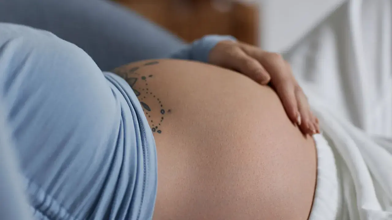 Dłoń spoczywa na **twardym brzuchu w ciąży**, ozdobionym delikatnym tatuażem.