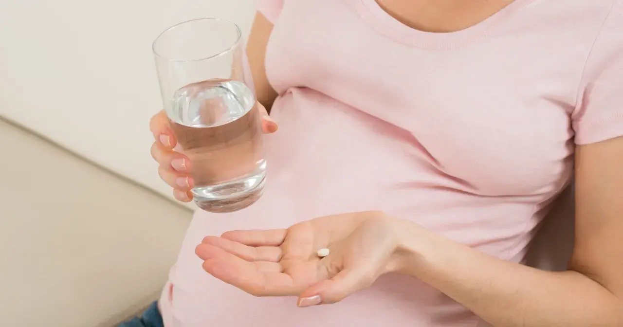 Kobieta w ciąży trzyma tabletkę i szklankę wody, szukając odpowiedzi na pytanie: co na ból głowy w ciąży.