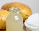 Naturalny sok z ziemniaka w buteleczce, obok surowych ziemniaków i wacików. Idealny do domowych zabiegów pielęgnacyjnych.
