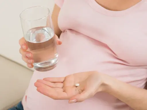 Kobieta w ciąży trzyma tabletkę i szklankę wody, szukając odpowiedzi na pytanie: co na ból głowy w ciąży.