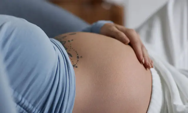 Dłoń spoczywa na **twardym brzuchu w ciąży**, ozdobionym delikatnym tatuażem.
