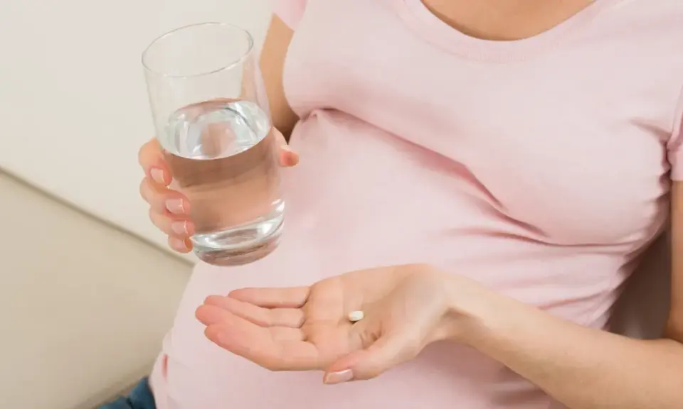 Kobieta w ciąży trzyma tabletkę i szklankę wody, szukając odpowiedzi na pytanie: co na ból głowy w ciąży.